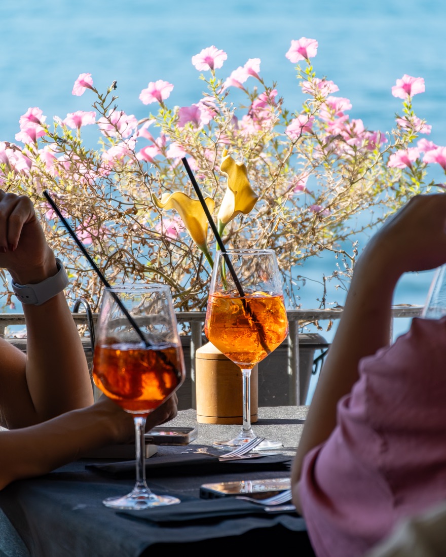 Two spritzes on a café table by the water in Italy, with pink flowers behind
